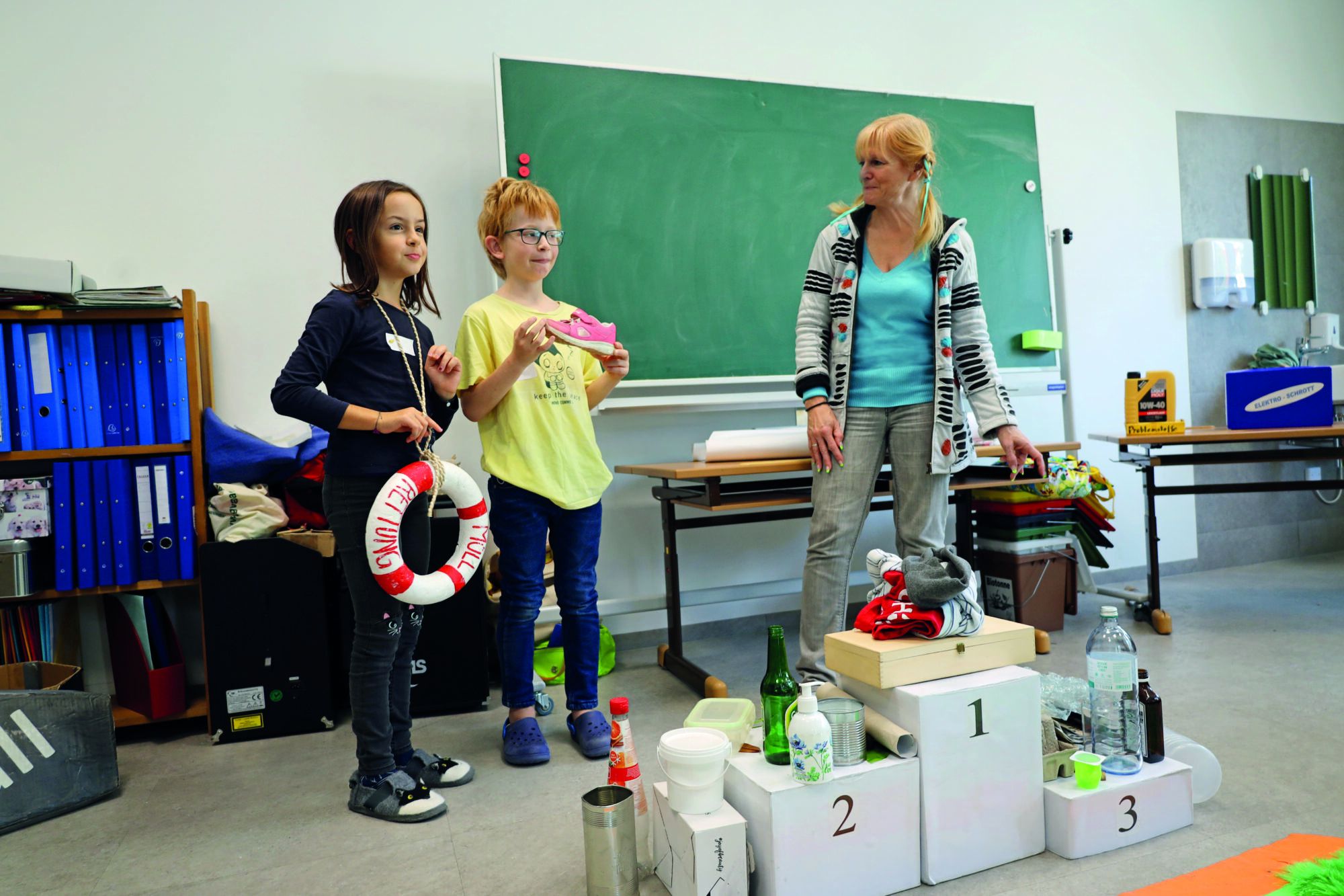 Zwei Kinder im Volksschulalter stehen neben einer bloden Frau mit geflochtenen Zu00f6pfen und einer Schleife im Haar. Davor ein Siegespodest mit Abfu00e4llen darauf und im Hintergrund eine Tafel und ein Regal mit blauen Ordnern. 