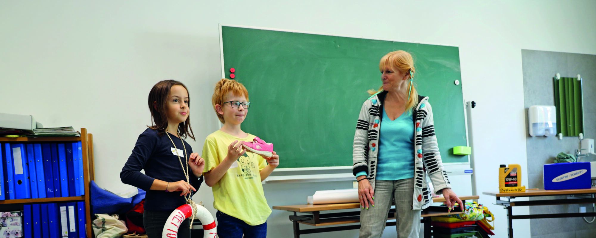 Zwei Kinder im Volksschulalter stehen neben einer bloden Frau mit geflochtenen Zu00f6pfen und einer Schleife im Haar. Davor ein Siegespodest mit Abfu00e4llen darauf und im Hintergrund eine Tafel und ein Regal mit blauen Ordnern. 