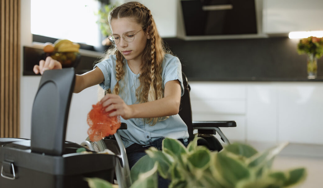 Auf dem Bild ist ein Mädchen zu sehen, dass im Rollstuhl sitzt und ein oranges Obstnetz in einen Abfallbehälter wirft. Im Vordergrund ist auch eine Pflanze zu sehen.