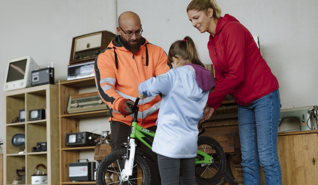 Ein kleines M&auml;dchen bekommt in einem ReUse and Repair Shop ein abgegebenes Fahrrad von dem Mitarbeiter in die Hand gedr&uuml;ckt.